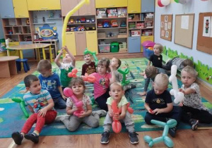 Dzieci siedzą na dywanie trzymając w rękach zwierzątka wykonane z balonów